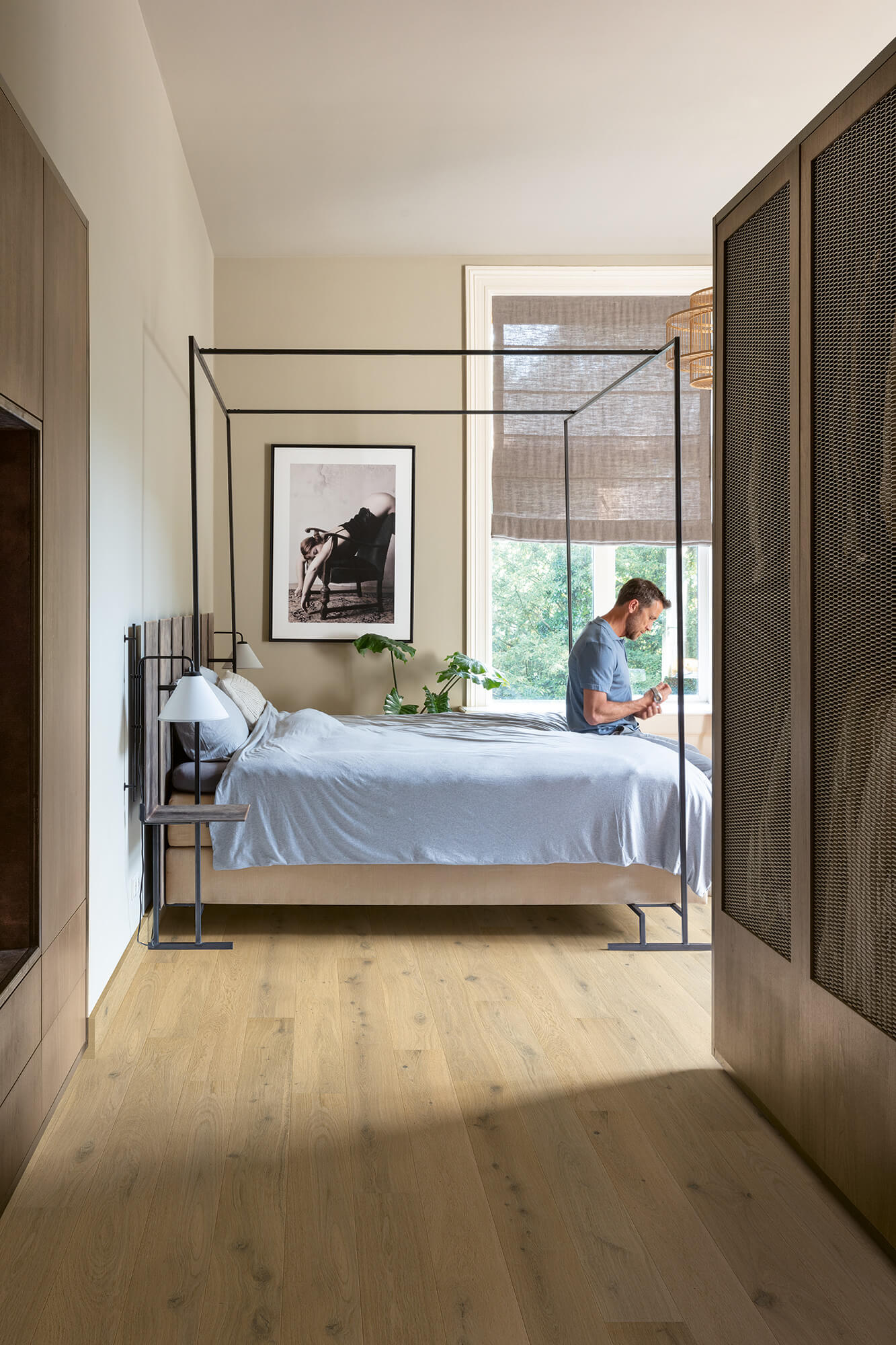 Modern bedroom with a glass canopy bed, wooden floor, and a person reading a book.