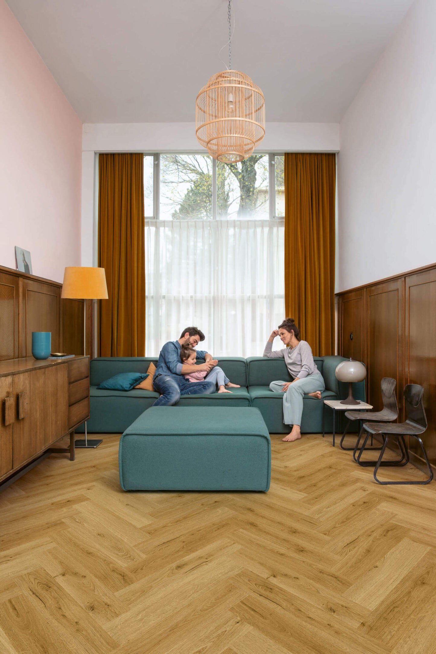 Family sitting on a teal sectional sofa in a modern living room with wooden floors and large windows.