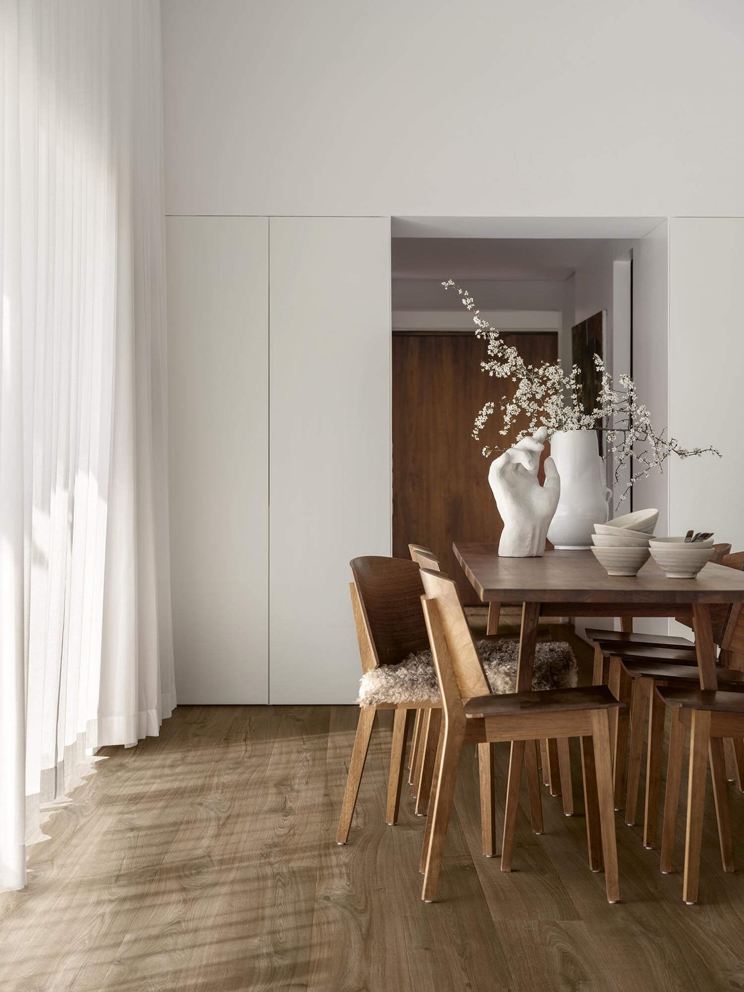 Dining area with wooden table and chairs, white vase with branches, and light-colored walls.