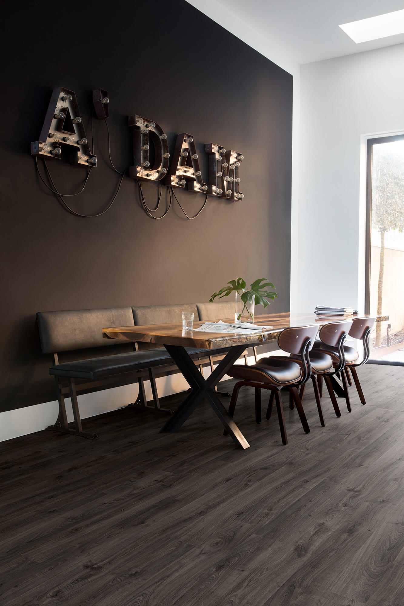 Modern dining area with wooden table and chairs against a dark wall with 'ADAM' letters.