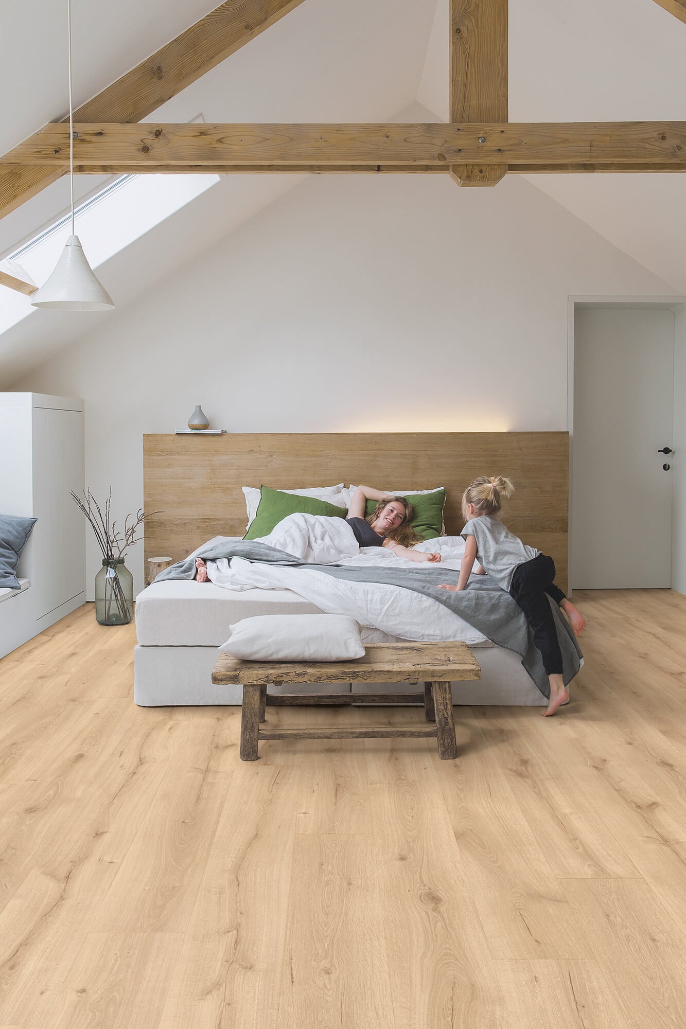 Two people lying on a bed in a modern bedroom with wooden flooring and exposed beams.