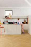 Two people in a modern kitchen with light-colored cabinets and wooden flooring.