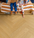 Children sitting on a bed with striped bedding, looking down at a wooden floor.