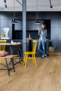 Woman standing in a modern kitchen with yellow chairs and wooden flooring.
