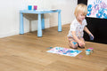 Child painting on a wooden floor with a small table and cups in the background.