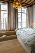 Modern bedroom with large windows, wooden floor, and a bed with light-colored bedding.
