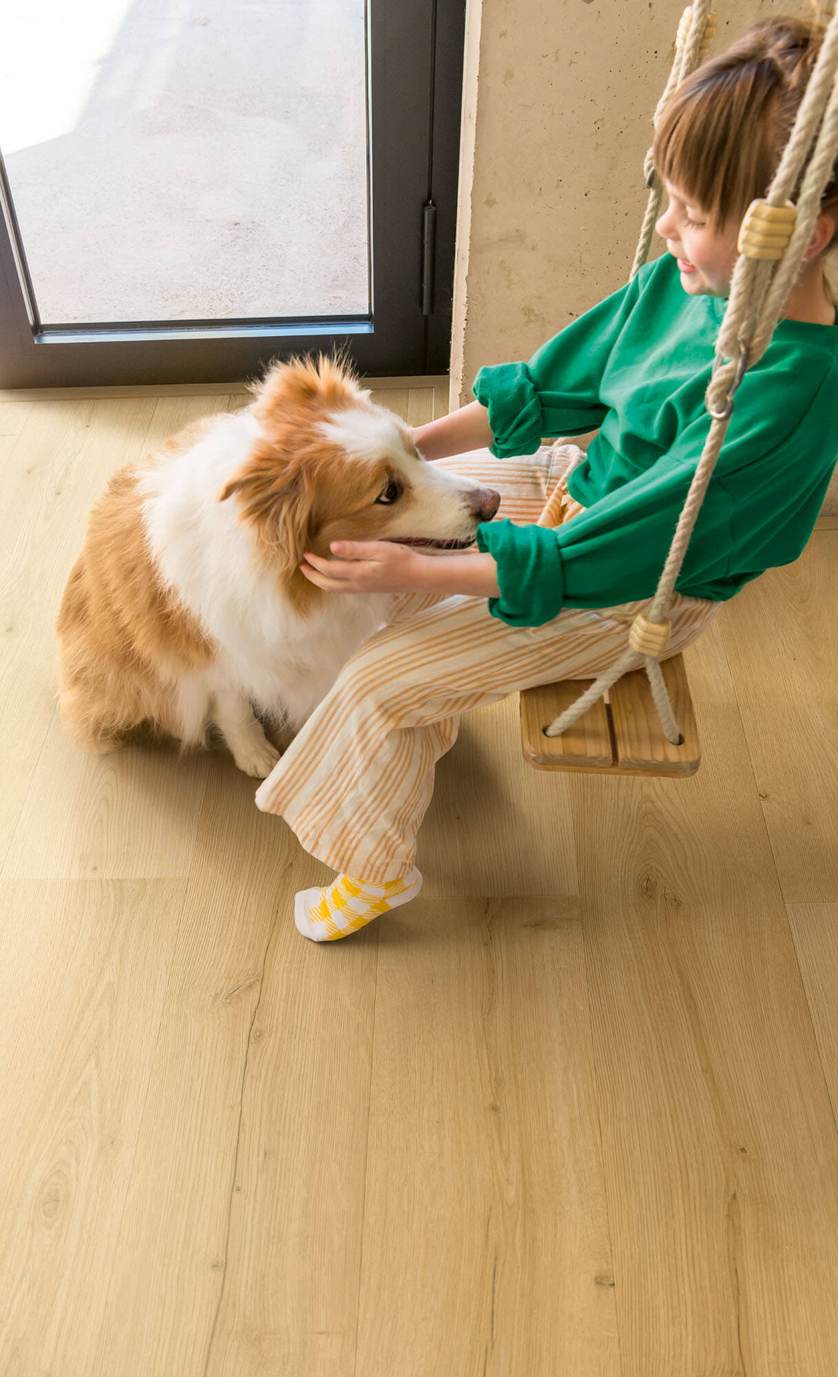 Child sitting on a swing with a dog on a wooden floor.