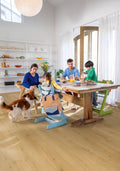 Family with children and a dog gathered around a wooden dining table in a bright room.