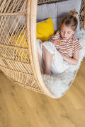 Child sitting in a wicker egg chair on a wooden floor with a yellow pillow and gray cushion.