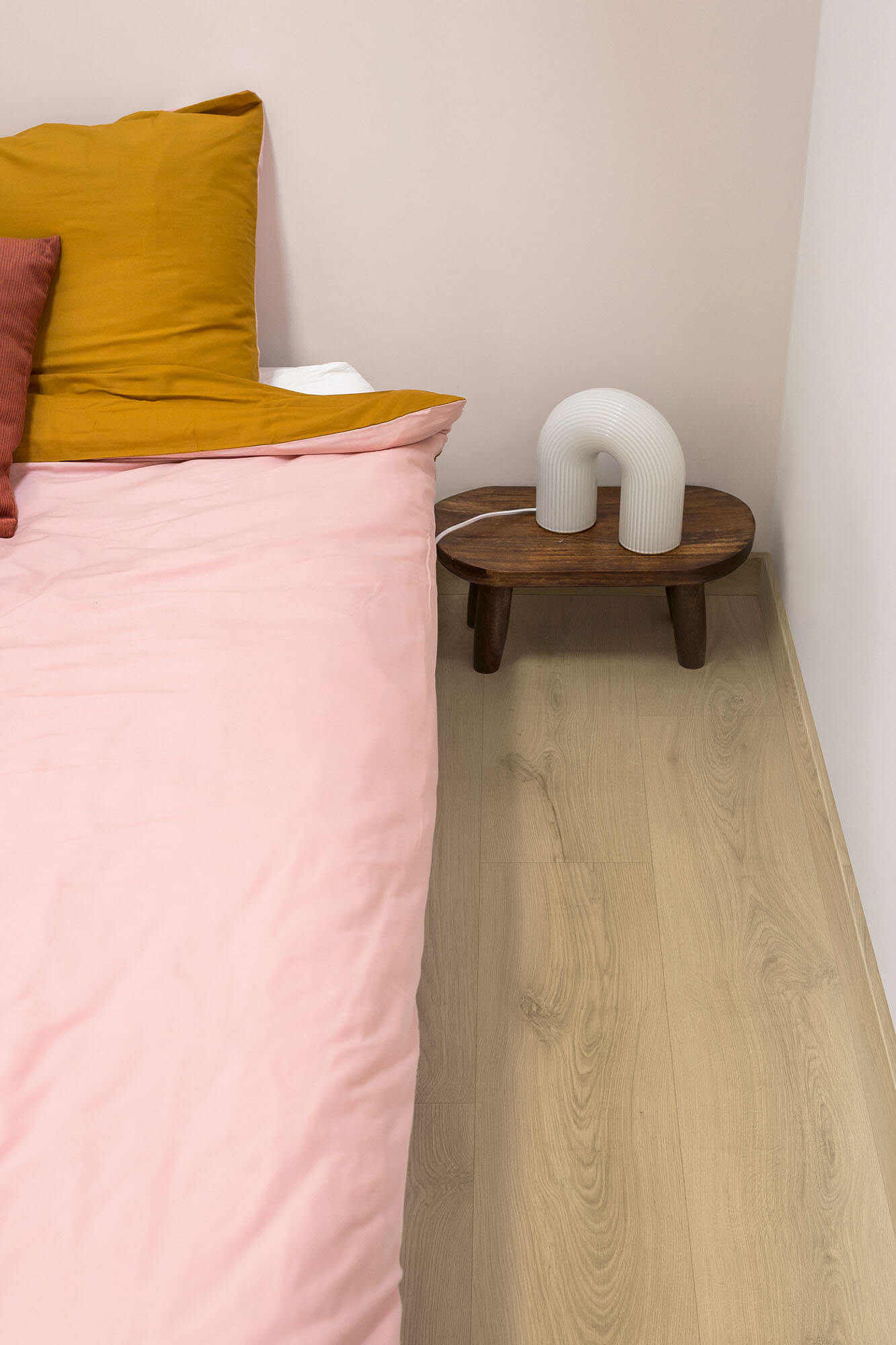 Bed with pink bedding and yellow pillow, wooden side table with white cylindrical object, light wood floor.