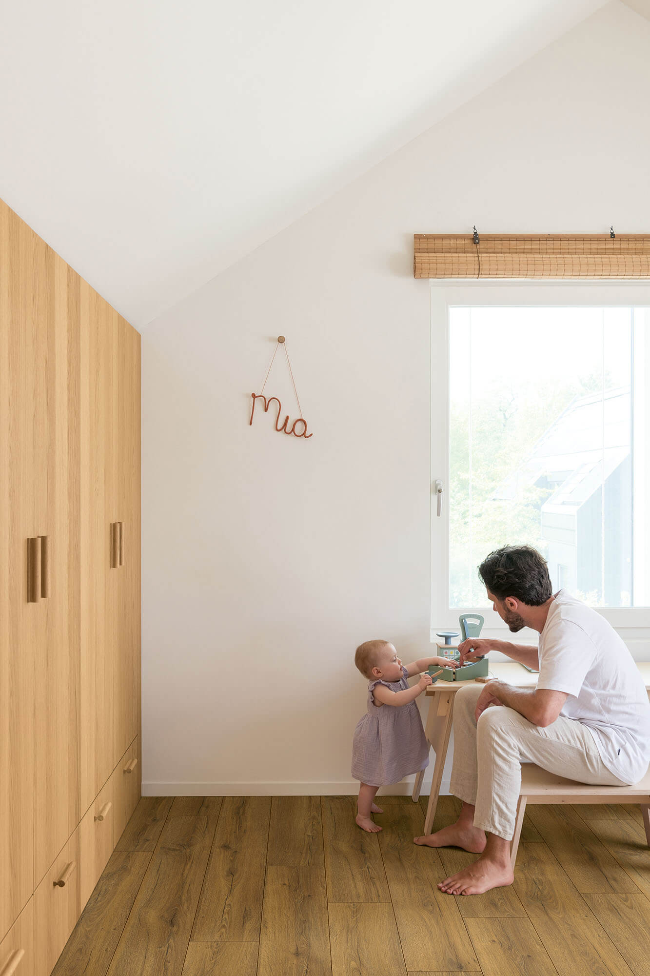 Man and child sitting at a small table in a room with wooden flooring and a window.