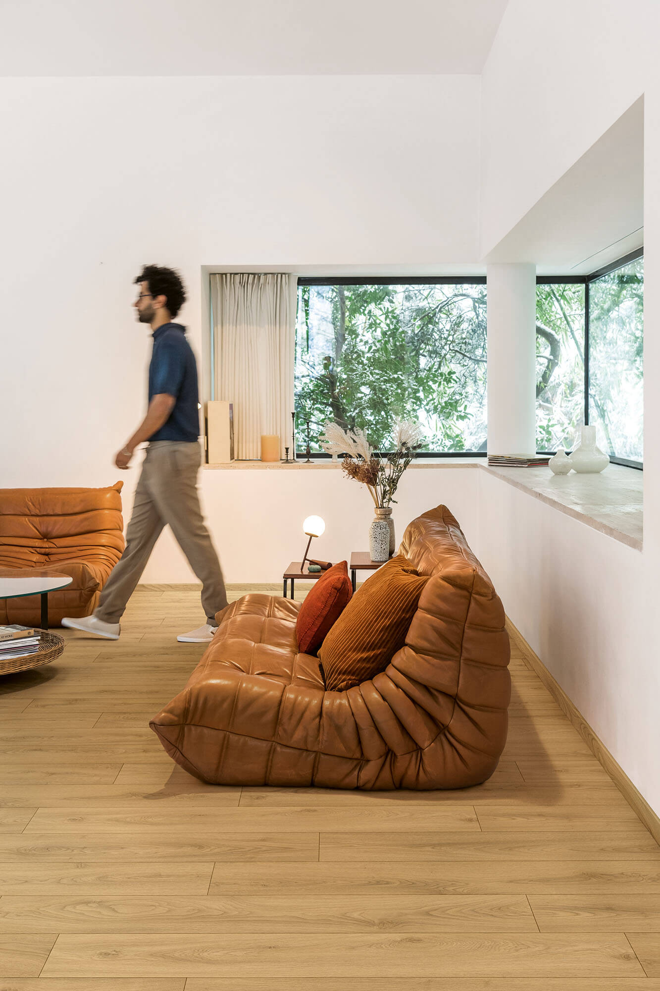 Man walking past a large brown bean bag chair in a modern living room with large windows.