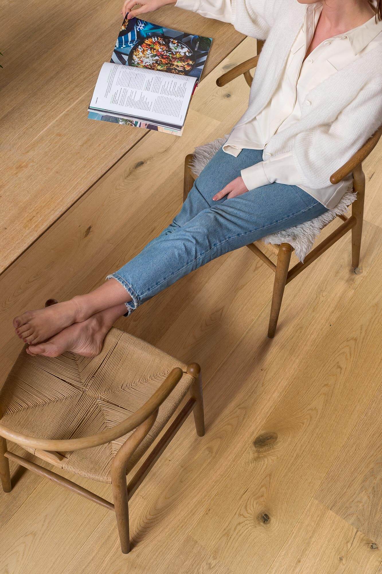 Person sitting on a wooden chair with a footstool, holding a magazine on a wooden floor.