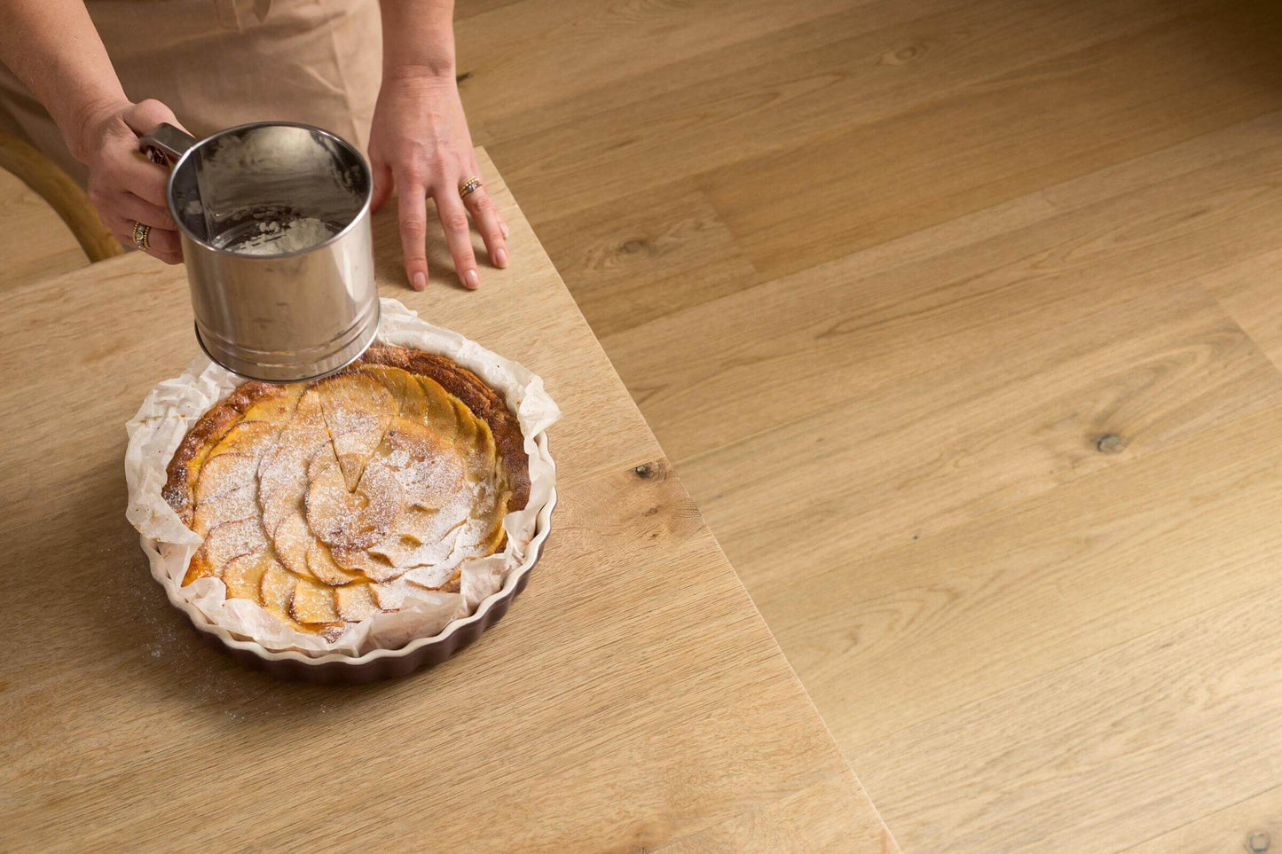 Person dusting an apple pie with flour using a sifter on a wooden table.