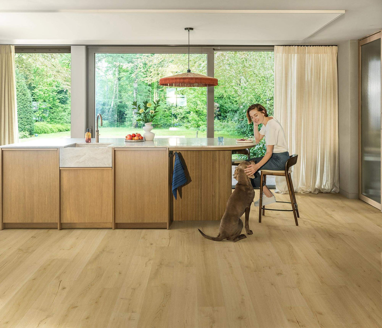 Woman sitting on a stool in a modern kitchen with a dog, surrounded by large windows with a view of greenery.