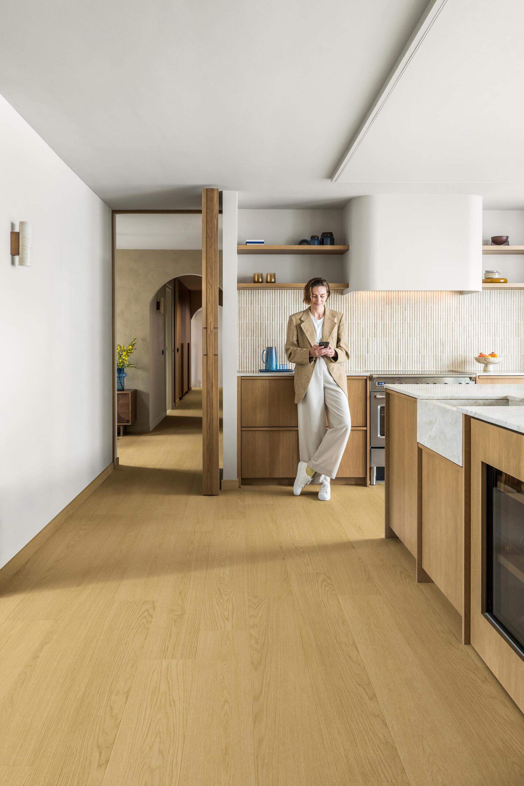 A woman in a beige blazer and white pants stands in a modern kitchen with light wood flooring and cabinetry, holding a phone.