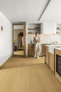 A woman in a beige blazer and white pants stands in a modern kitchen with light wood flooring and cabinetry, holding a phone.