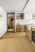 A modern kitchen interior with light oak wood flooring, light wood cabinetry, white walls, and a tiled backsplash. An open doorway on the left reveals an arched hallway in the distance.