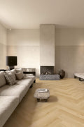A modern living room with a light beige sectional sofa, a low stone coffee table, a black floor lamp, and a central fireplace with a textured surround, set on light herringbone wood flooring.