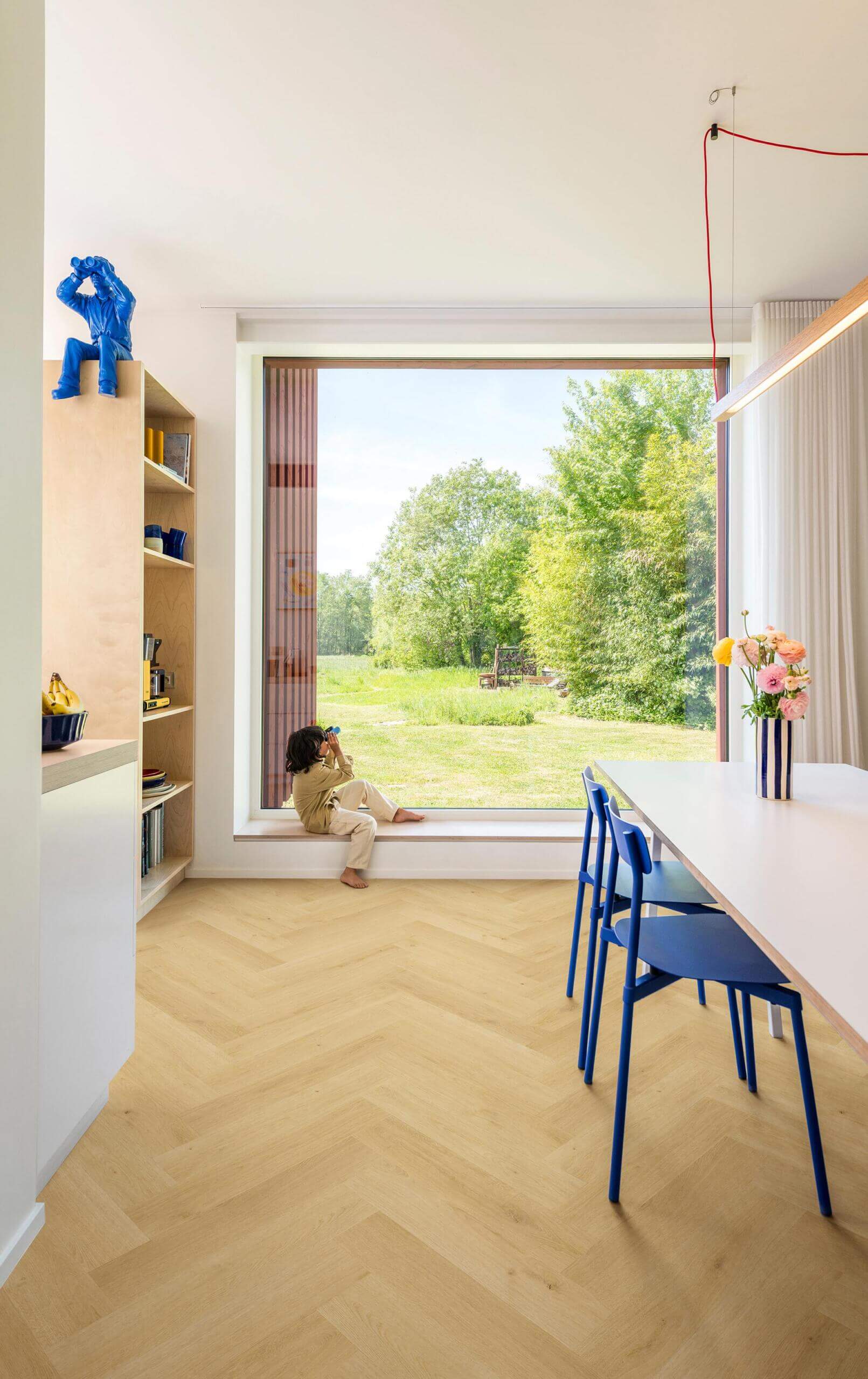 Modern interior with a large window view of greenery, a child sitting on the windowsill, and a desk with chairs.