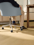 A person seated in a light blue upholstered office chair with a chrome five-star base and black casters, shown in a sunlit home office with light wood flooring and a desk.