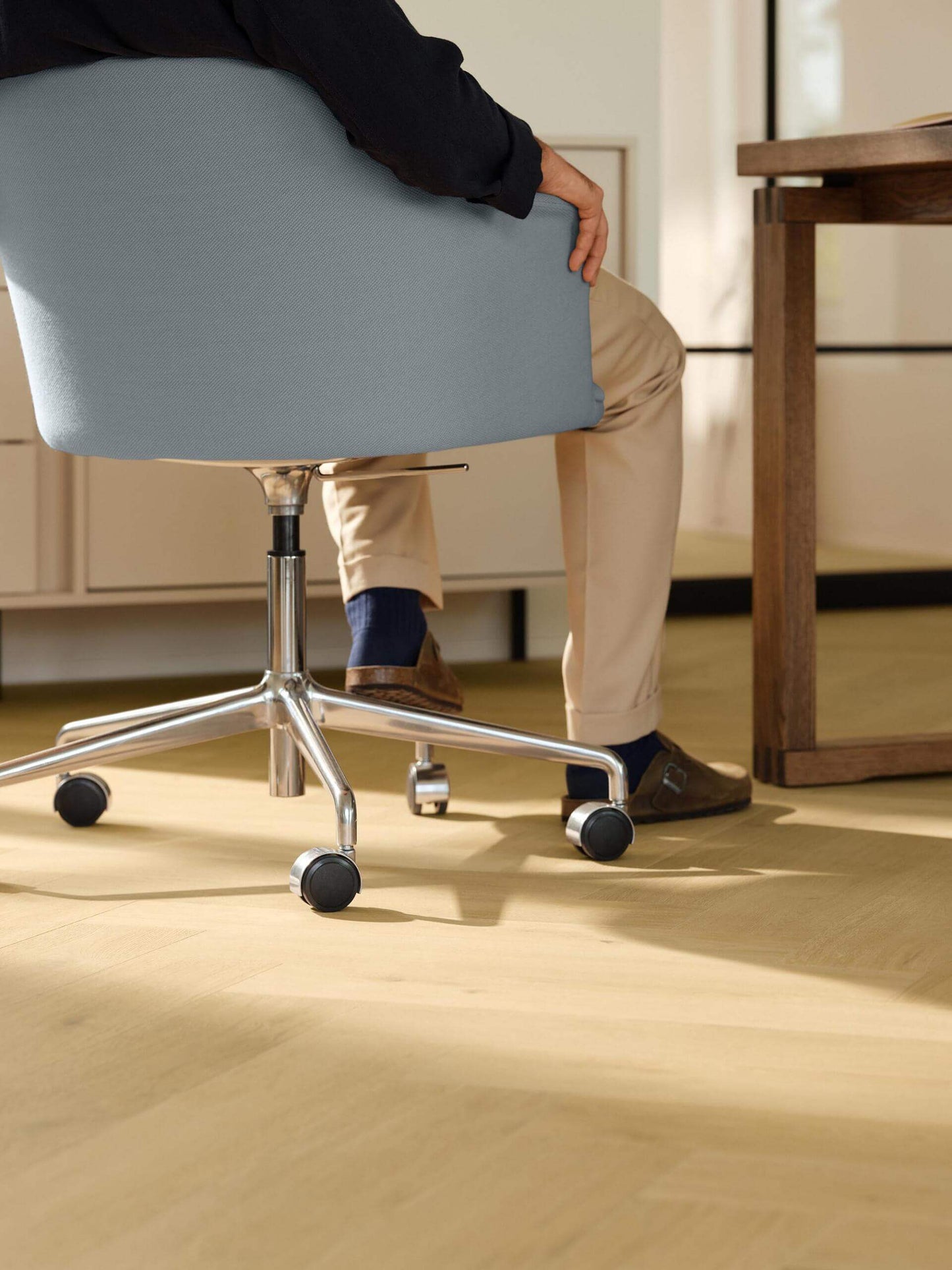 A person seated in a light blue upholstered office chair with a chrome five-star base and black casters, shown in a sunlit home office with light wood flooring and a desk.