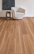 Light brown wood plank flooring shown in a modern living room setting with a white armchair, a small rustic side table, and a white paneled wall.