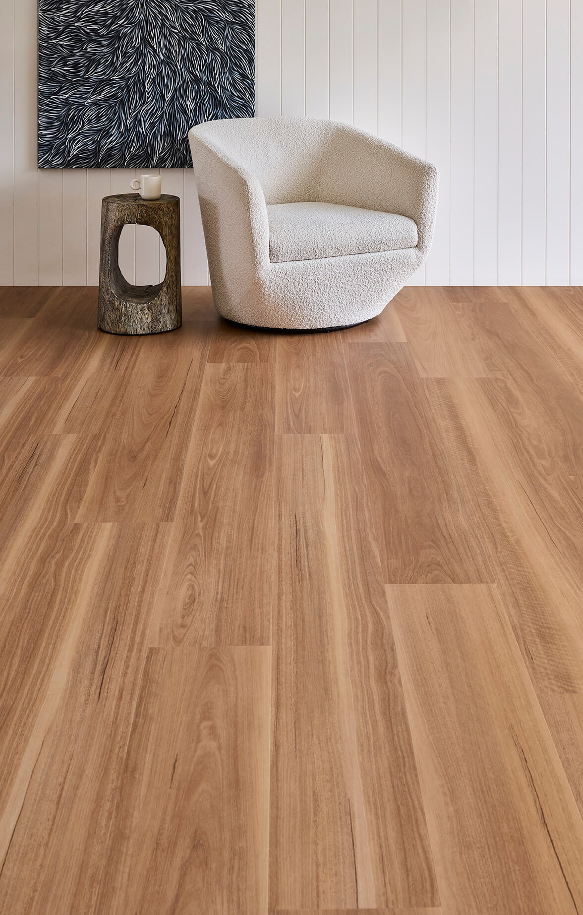 Light brown wood plank flooring shown in a modern living room setting with a white armchair, a small rustic side table, and a white paneled wall.