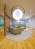 A modern interior lounge area with a curved beige sofa, matching ottoman, low wooden coffee table, and a large circular rug on a light wood floor. The space features a sculptural circular ceiling light, wood slat walls, and glass windows in the background.