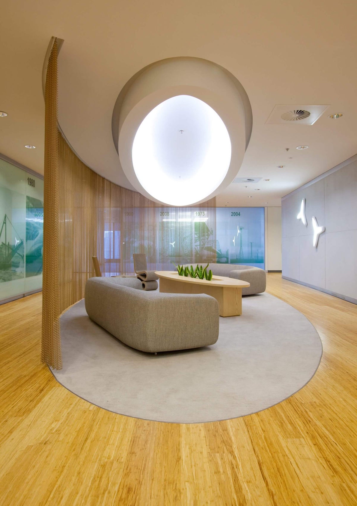 A modern interior lounge area with a curved beige sofa, matching ottoman, low wooden coffee table, and a large circular rug on a light wood floor. The space features a sculptural circular ceiling light, wood slat walls, and glass windows in the background.