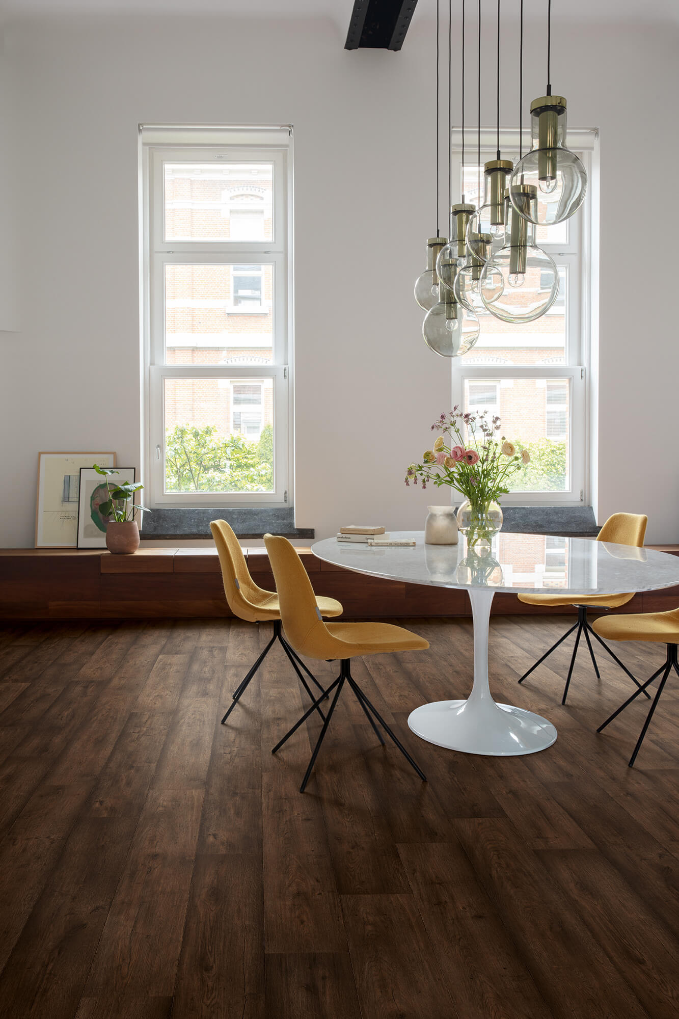 Modern dining room with round white table and yellow chairs, large windows, and pendant lights.