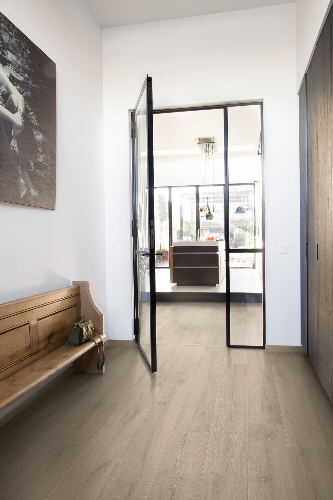 Modern interior with a glass door leading to a kitchen, wooden bench on the left, and artwork on the wall.