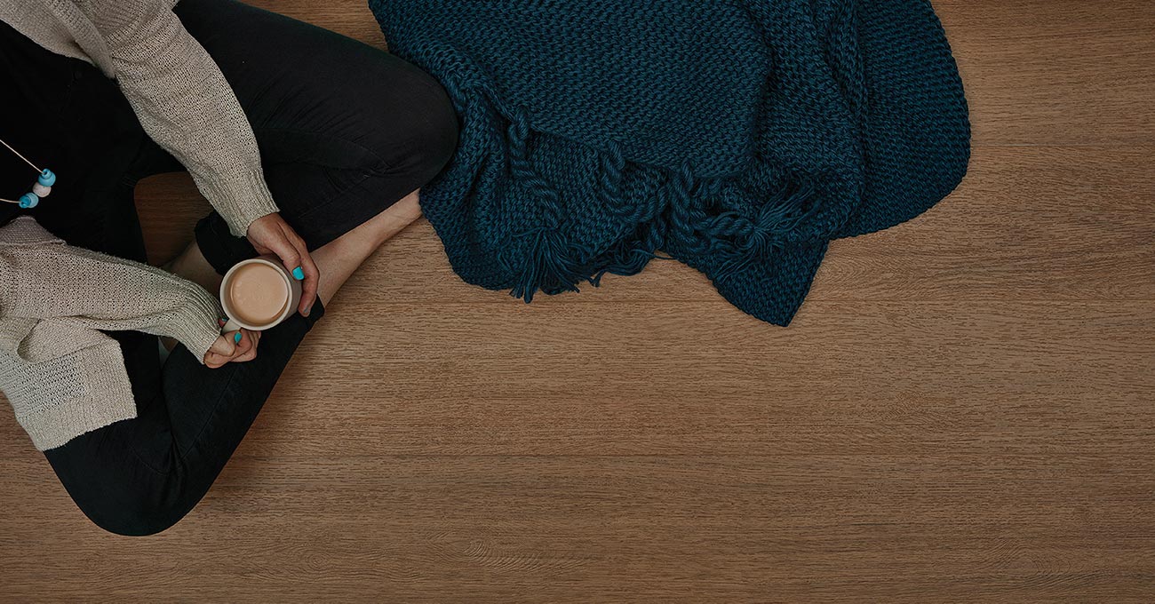 Person sitting on a wooden floor holding a cup, with a blue knitted blanket nearby.