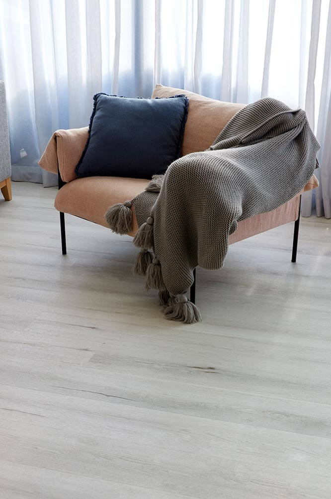 Pink sofa with a gray blanket and blue pillow in a room with light-colored wooden flooring and curtains.