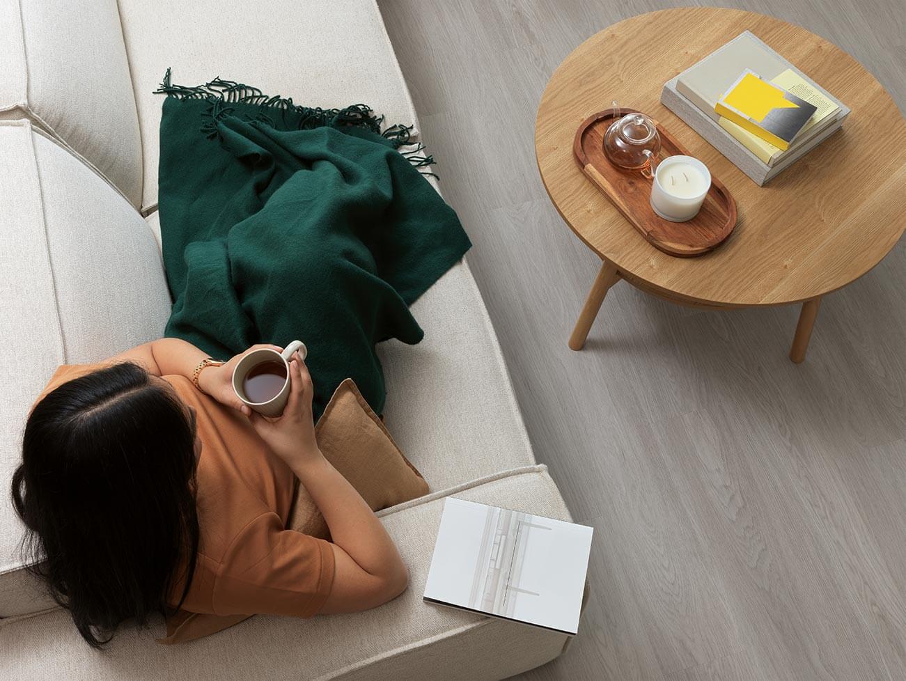 Person lying on a couch with a green blanket, holding a mug, next to a wooden coffee table with a candle and books.